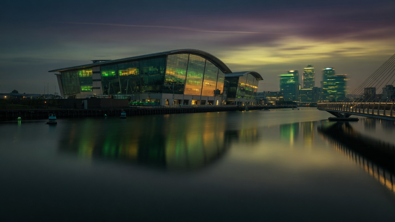Dusk view of ExCeL London riverside architecture reflecting green and gold lights on calm Docklands water